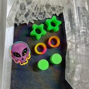 May include: A set of green star-shaped and round acrylic ear gauges, a pair of green acrylic ear plugs, and a holographic sticker of an alien with a galaxy print.