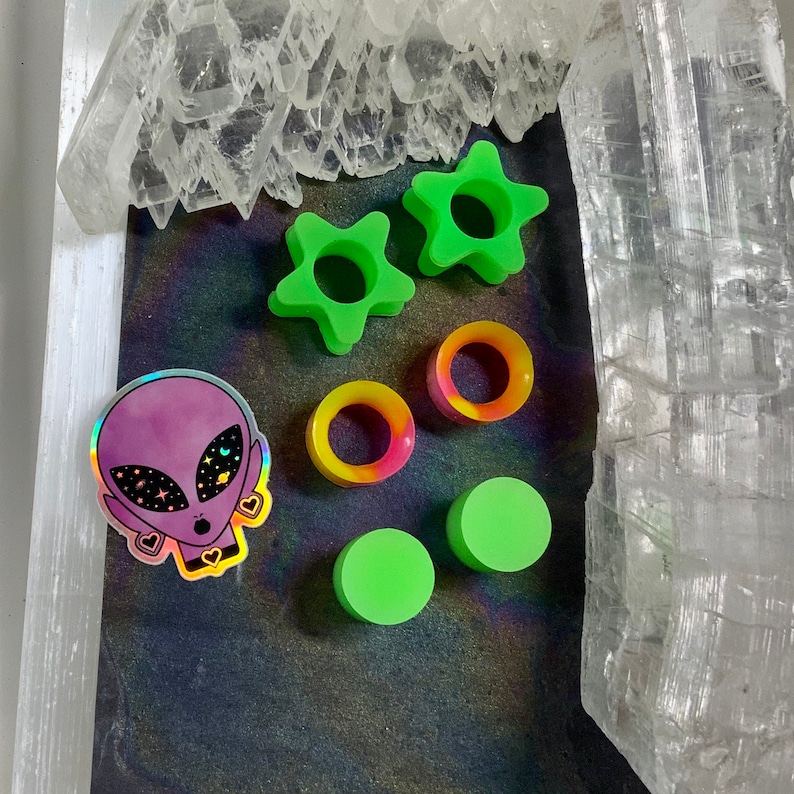 May include: A set of green star-shaped and round acrylic ear gauges, a pair of green acrylic ear plugs, and a holographic sticker of an alien with a galaxy print.