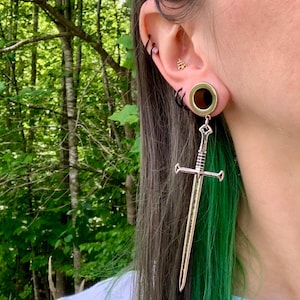 May include: A silver-toned sword-shaped earring pendant hanging from a circular ear gauge. The sword features a detailed hilt, crossguard, and blade. The ear gauge is metallic with a dark centre, and the ear also displays a small gold symbol stud.