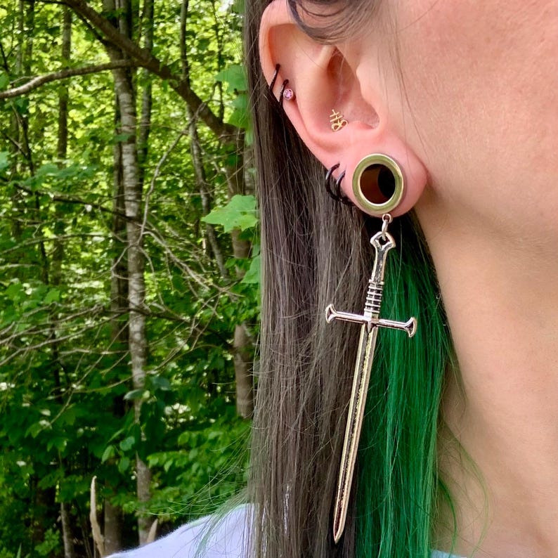 May include: A silver-toned sword-shaped earring pendant hanging from a circular ear gauge. The sword features a detailed hilt, crossguard, and blade. The ear gauge is metallic with a dark centre, and the ear also displays a small gold symbol stud.