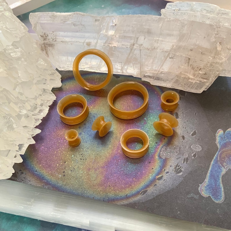 May include: A set of eight brown silicone ear gauges of varying sizes, displayed on a black surface with iridescent rainbow patterns.