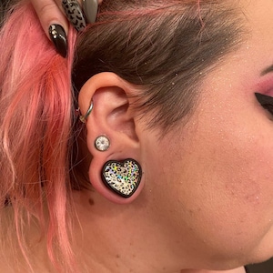 May include: A close-up of a person's ear with a black heart-shaped ear gauge with a leopard print design. The ear gauge is worn in the lower lobe of the ear.