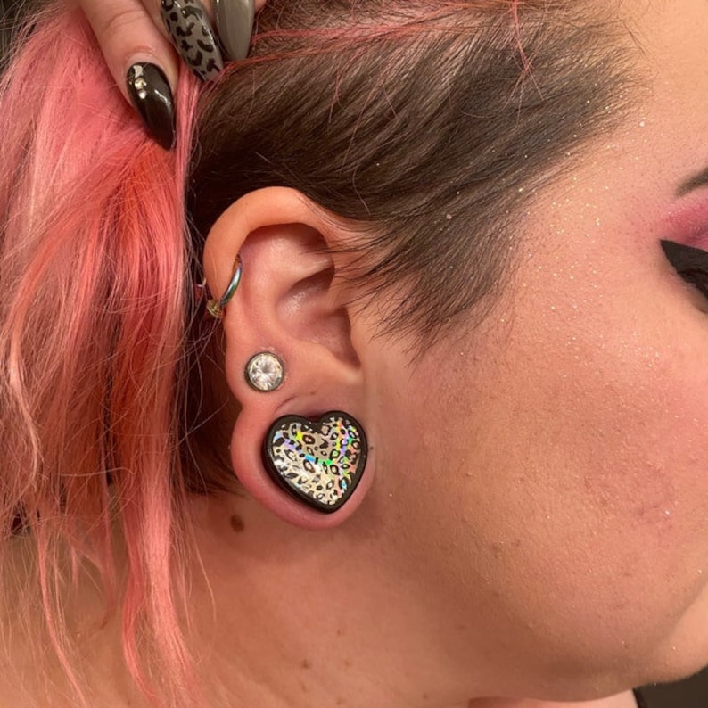 May include: A close-up of a person's ear with a black heart-shaped ear gauge with a leopard print design. The ear gauge is worn in the lower lobe of the ear.