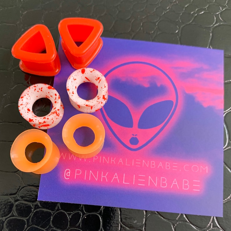 May include: Set of six silicone ear gauges in various colors and designs, including orange, white with red splatter, and red triangle shapes. The gauges are displayed on a purple background with an alien face graphic and the text "www.pinkalienbabe.com @pinkalienbabe".
