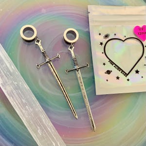 May include: A pair of silver-toned sword-shaped ear plugs with circular tunnels. The packaging is a clear pouch with a heart-shaped cutout, star and planet graphics, and the text "PINK ALIEN BABE" with a pink sticker indicating 1/2" 12mm.