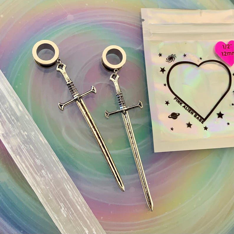 May include: A pair of silver-toned sword-shaped ear plugs with circular tunnels. The packaging is a clear pouch with a heart-shaped cutout, star and planet graphics, and the text "PINK ALIEN BABE" with a pink sticker indicating 1/2" 12mm.