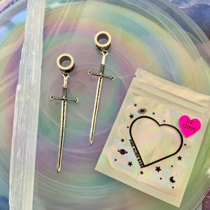 May include: A pair of silver-toned sword-shaped ear plugs with circular tunnels, measuring 12mm. The packaging is a clear plastic bag with a heart-shaped cutout, featuring the text PINK ALIEN BABE and a pink sticker stating 1/2 inch 12mm.