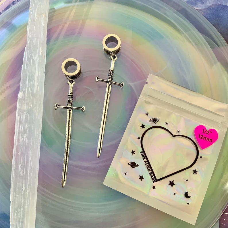 May include: A pair of silver-toned sword-shaped ear plugs with circular tunnels, measuring 12mm. The packaging is a clear plastic bag with a heart-shaped cutout, featuring the text PINK ALIEN BABE and a pink sticker stating 1/2 inch 12mm.