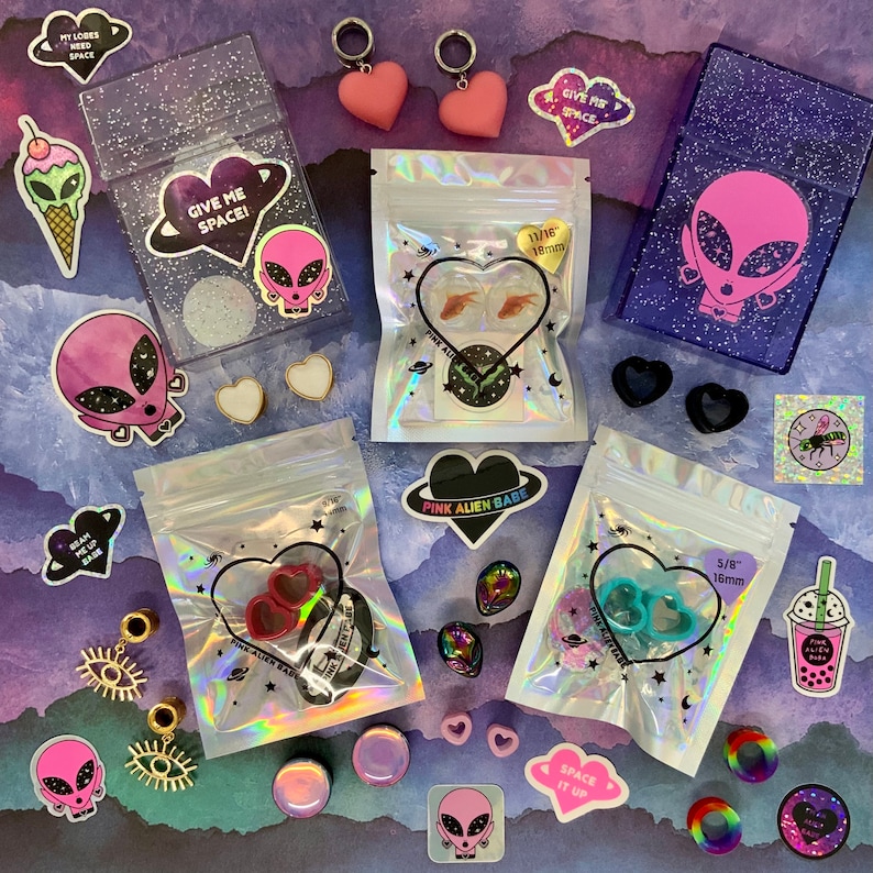 May include: A collection of colorful, glittery, and holographic packaging for jewelry and accessories. The packaging features alien and heart designs with the text "Give Me Space!" and "Pink Alien Babe".