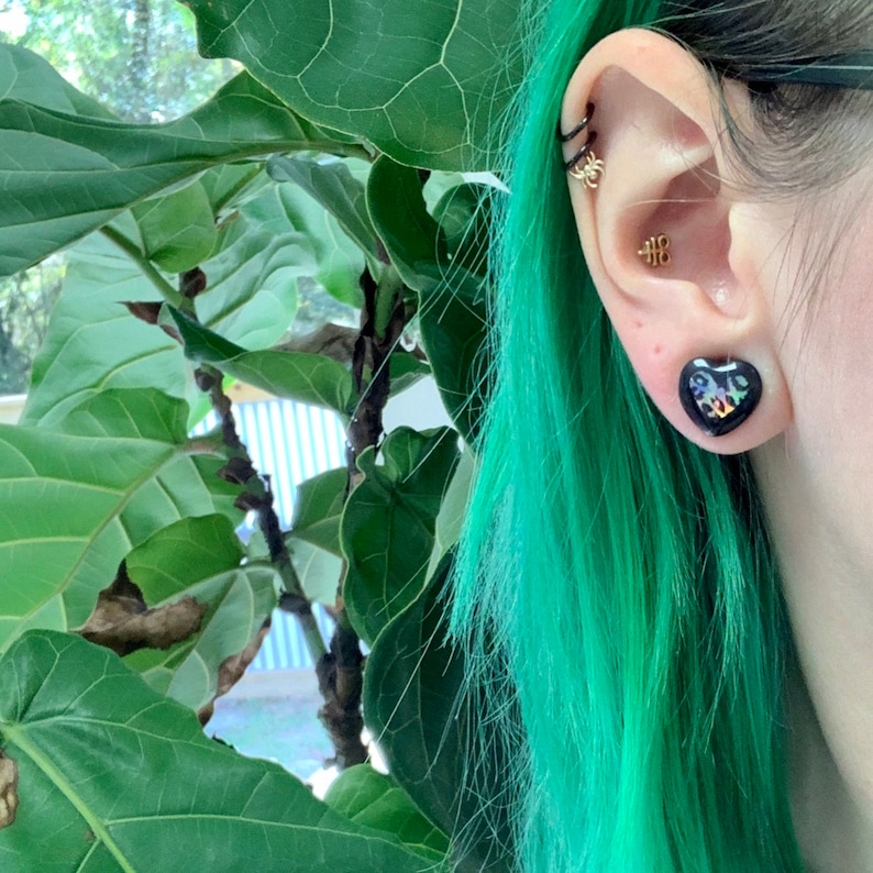 May include: A person with teal green hair wearing a black heart-shaped earring. The earring has a glittery, iridescent finish.