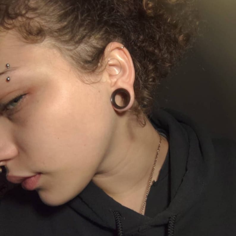 May include: A close-up of a person's face with a black hoop earring in their right ear. The person has short, curly brown hair and is wearing a black hoodie.