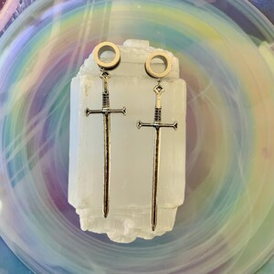 May include: A pair of silver-toned metal ear plugs featuring dangling sword charms. Each plug has a circular tunnel design at the top, connected to a long, slender sword pendant with a detailed hilt, crossguard, and pointed blade tip.