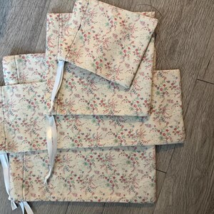 Puede incluir: Un conjunto de art&iacute;culos de tela color crema con un estampado floral en rosa, rojo y verde. Los art&iacute;culos parecen estar doblados y atados con cintas blancas. La tela tiene una est&eacute;tica delicada y vintage.