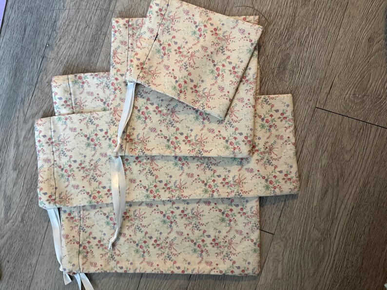 Puede incluir: Un conjunto de art&iacute;culos de tela color crema con un estampado floral en rosa, rojo y verde. Los art&iacute;culos parecen estar doblados y atados con cintas blancas. La tela tiene una est&eacute;tica delicada y vintage.