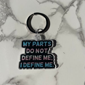 Puede incluir: Un llavero de acrílico negro con el texto "Mis partes no me definen; yo me defino a mí mismo." El texto está en blanco con un diseño de bandera del orgullo transgénero rosa y azul.