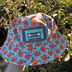 May include: A bucket hat with a light blue background and a pattern of orange flowers. A rectangular patch with a teal cassette tape design is sewn on the front. The hat is made of fabric and is suitable for outdoor wear.