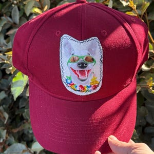 Puede incluir: Gorra de béisbol burdeos con un parche que muestra un perro sonriente con gafas de sol. El parche tiene un fondo blanco y un diseño floral colorido. La gorra está hecha de una tela resistente y tiene una visera curva.