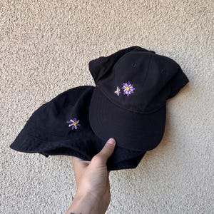 May include: Two black hats, a baseball cap and a bucket hat, each embroidered with a small purple and white daisy. The hats are made of a soft fabric and are suitable for casual wear.