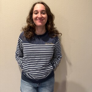 May include: A navy blue and white striped sweatshirt with the embroidered text "They/Them" in gold thread. The sweatshirt is paired with denim shorts.
