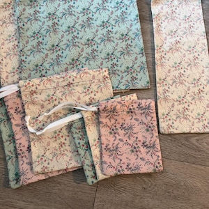 Puede incluir: Una colecci&oacute;n de bolsas de tela de varios tama&ntilde;os, con un estampado floral con detalles en rosa, azul y rojo sobre un fondo crema y pastel. Algunas bolsas tienen cierres de cord&oacute;n blancos. La tela tiene una est&eacute;tica vintage.