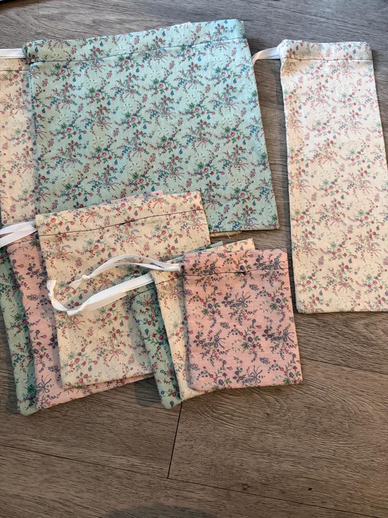 Puede incluir: Una colecci&oacute;n de bolsas de tela de varios tama&ntilde;os, con un estampado floral con detalles en rosa, azul y rojo sobre un fondo crema y pastel. Algunas bolsas tienen cierres de cord&oacute;n blancos. La tela tiene una est&eacute;tica vintage.