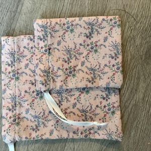 Puede incluir: Dos bolsas de tela rosa con un estampado floral de flores y lazos azules, morados y rojos. Las bolsas son rectangulares y tienen un cierre de cinta blanca. La tela tiene un fondo rosa claro.