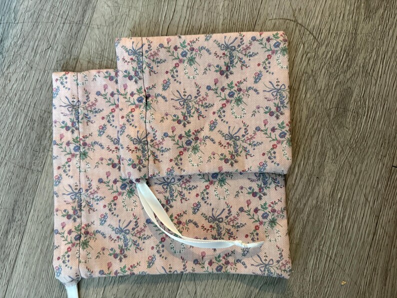 Puede incluir: Dos bolsas de tela rosa con un estampado floral de flores y lazos azules, morados y rojos. Las bolsas son rectangulares y tienen un cierre de cinta blanca. La tela tiene un fondo rosa claro.