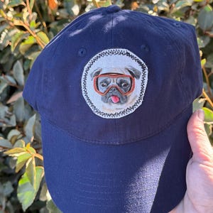 May include: Navy blue baseball cap with a circular patch featuring a pug wearing orange goggles. The pug has its tongue sticking out. The cap is made of a durable fabric and has a curved brim.