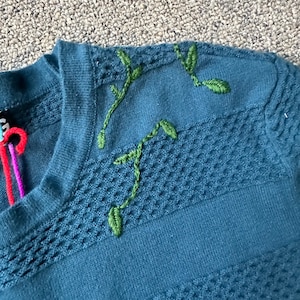 Peut inclure: Un pull en tricot bleu foncé avec un motif de vigne brodé en vert sur l'épaule.