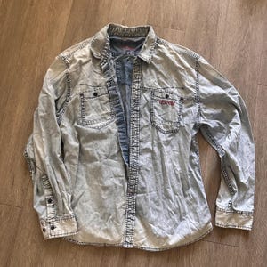 May include: A light blue denim shirt with long sleeves and a button-down front. The shirt has a red embroidered logo that says "DEFRIM" on the left chest pocket.