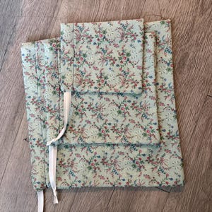 Puede incluir: Un conjunto de tres bolsas de tela con un estampado floral en tonos rosa, azul y verde sobre un fondo verde claro. Las bolsas est&aacute;n atadas con cintas blancas. La tela tiene una textura acolchada.