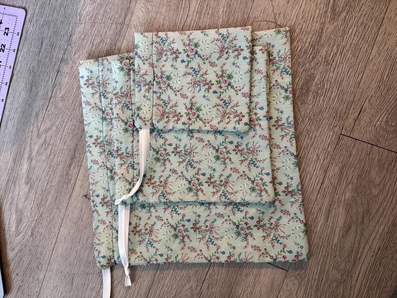 Puede incluir: Un conjunto de tres bolsas de tela con un estampado floral en tonos rosa, azul y verde sobre un fondo verde claro. Las bolsas est&aacute;n atadas con cintas blancas. La tela tiene una textura acolchada.