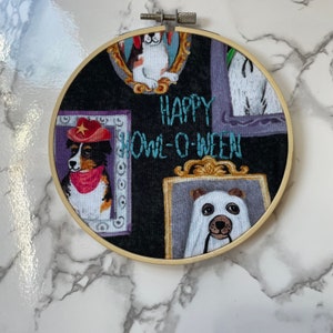 May include: A black fabric hoop with a gold frame featuring a variety of embroidered images of dogs and cats in picture frames. The text "HAPPY HOWL-O-WEEN" is embroidered in teal thread.