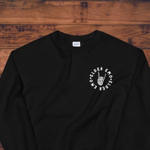 Pode incluir: Camisola de moletom preta com um gráfico circular branco que apresenta uma mão de esqueleto fazendo o sinal de rock and roll. O texto "EMO ELDER EMO ELDER" está escrito ao redor do círculo.