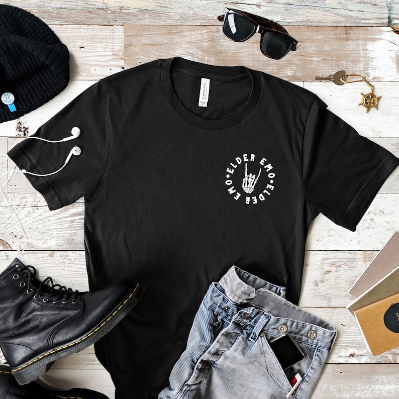 May include: Black t-shirt with the text "Elder Emo" in a circle around a skeleton hand making a rock and roll gesture. The shirt is paired with blue jeans, black boots, a black beanie, and sunglasses.