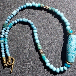 May include: A blue and turquoise beaded necklace with a large, patterned turquoise pendant. The necklace has a brass clasp.