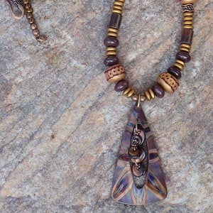 May include: A brown and gold necklace with a large, triangular pendant. The pendant is made of metal and has a geometric design. The necklace is made of beads and has a clasp closure.