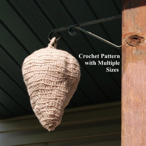 May include: A crocheted beige teardrop-shaped object hangs from a metal hook attached to a wooden post. The text "Crochet Pattern with Multiple Sizes" is visible below the object.