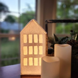 Może przedstawiać: Podświetlana, biała, filcowa dekoracja w kształcie domu z prostokątnymi oknami. Obok domu znajdują się dwie białe świece kolumnowe na baterie. Scena rozgrywa się na drewnianej powierzchni z rozmytym tłem.