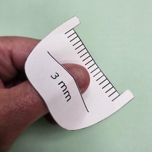 Op de afbeelding: Een wit papieren meetinstrument met zwarte markeringen en de tekst "3 mm". Het gereedschap wordt door een hand vastgehouden en bevindt zich tegen een lichtgroene achtergrond. Het gereedschap heeft een gebogen vorm.