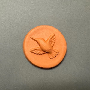 May include: A round, terracotta-colored ceramic piece with a raised hummingbird design. The bird is depicted in flight, wings outstretched, centered on the circular base. The background is a solid, neutral gray.