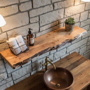 May include: A rustic bathroom vanity with a copper sink and a live-edge wooden shelf. The vanity has a wooden base and a copper faucet. A wire basket holds white towels. The wall is made of stone.