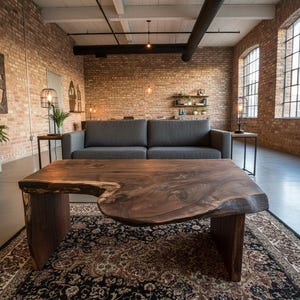 May include: A living room featuring a large, dark wood coffee table with a live edge design. A gray sofa sits against a brick wall, with a patterned rug beneath the table. The room has large windows and potted plants.
