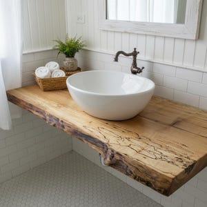 May include: A white ceramic vessel sink sits atop a natural live-edge wood countertop. The vanity features a dark bronze faucet, and a woven basket containing three white rolled towels and a small green potted plant rests on the wooden surface.