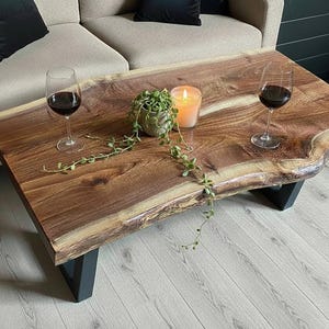 May include: A rectangular coffee table featuring a natural live edge wood slab top with a rich brown grain pattern, supported by two black metal U-shaped legs. The surface holds two clear glass wine stems, a small potted trailing plant, and a candle.