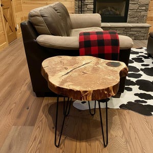 May include: A live edge wood side table with a natural light brown finish, featuring a prominent dark wood grain pattern, a central crack, and a dark circular inlay. The table is supported by three thin, black metal hairpin legs.