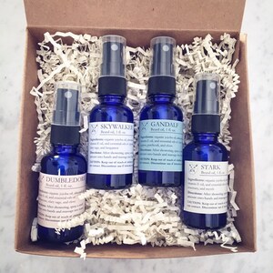 May include: Four blue glass bottles of beard oil, each with a black spray top, are arranged in a cardboard box with shredded paper. Each bottle has a different name: Dumbledore, Skywalker, Gandalf, and Stark. The bottles are 1 fl oz each.