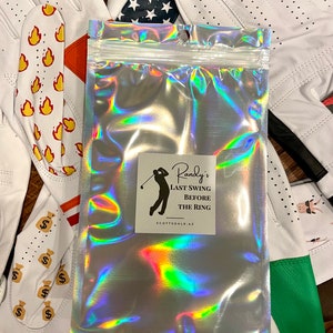May include: A silver holographic bag with a white label that says "Randy's Last Swing Before The Ring Scottsdale, AZ". The bag is surrounded by white golf gloves with different designs, including flames, dollar signs, stars, and shamrocks.