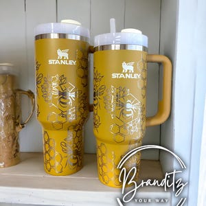 Op de afbeelding: Twee gele Stanley-bekers met witte deksels en zilveren bijen- en honingraatontwerpen. E&eacute;n beker heeft een handvat en een rietje. De bekers staan op een witte plank. De tekst "STANLEY" is zichtbaar.