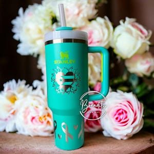 Op de afbeelding: Een turquoise Stanley-beker met een handvat en een rietje. De beker heeft een zilveren ontwerp van een hart, linten en het Stanley-logo. De handgreep en het deksel zijn licht turquoise van kleur. De achtergrond bevat witte en roze bloemen.